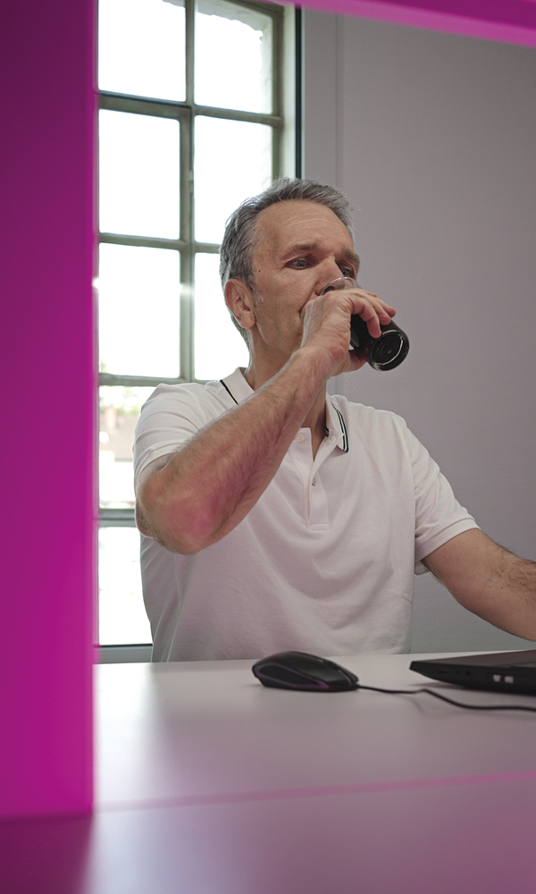 A middle-aged man in a white polo shirt sits in a pink-lit testing booth, drinking from a small glass while using a computer mouse; a laptop lies on the desk in front of him and a window is visible behind.