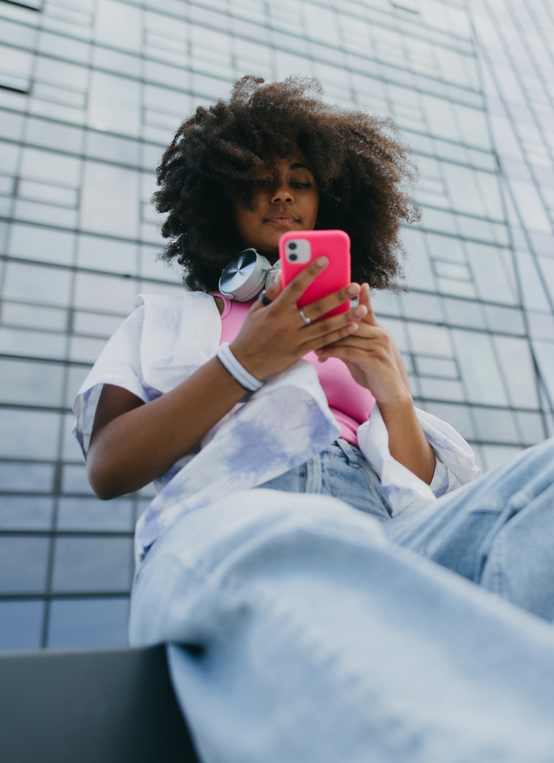 Young woman with curly afro hair, dressed casually in jeans, a pink tank top and open tie-dye shirt, wearing headphones around her neck, looking intently at her pink smartphone while seated against a modern glass building facade.