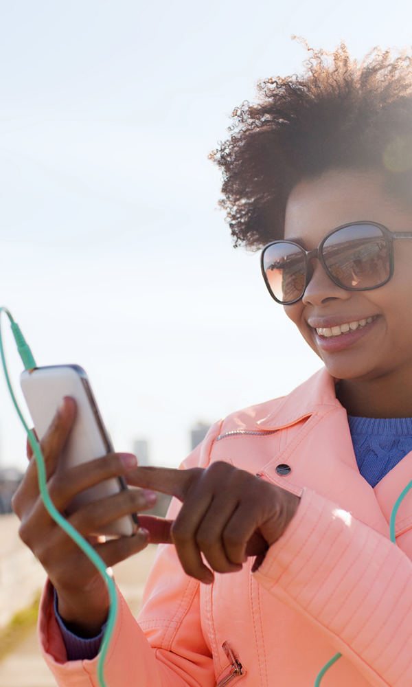Young woman with dark curly hair and large sunglasses, wearing a peach-colored jacket over a blue sweater, smiling as she taps on her smartphone connected to green earphones; a blurred cityscape and bright sky form the background.