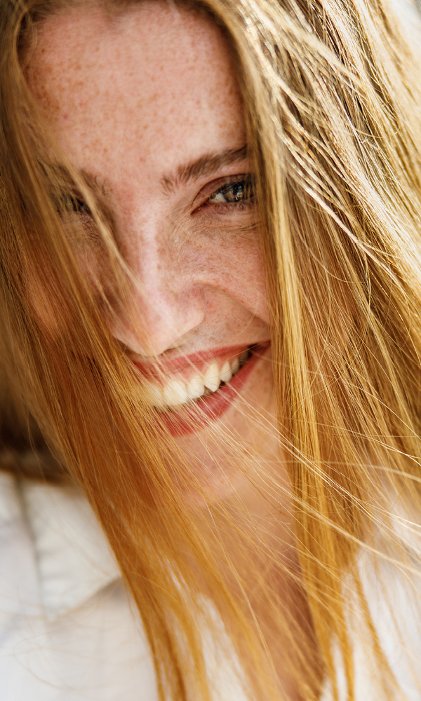 Close-up of a young red-haired woman with freckles, her long hair falling loosely across her face as she smiles broadly and looks slightly forward.