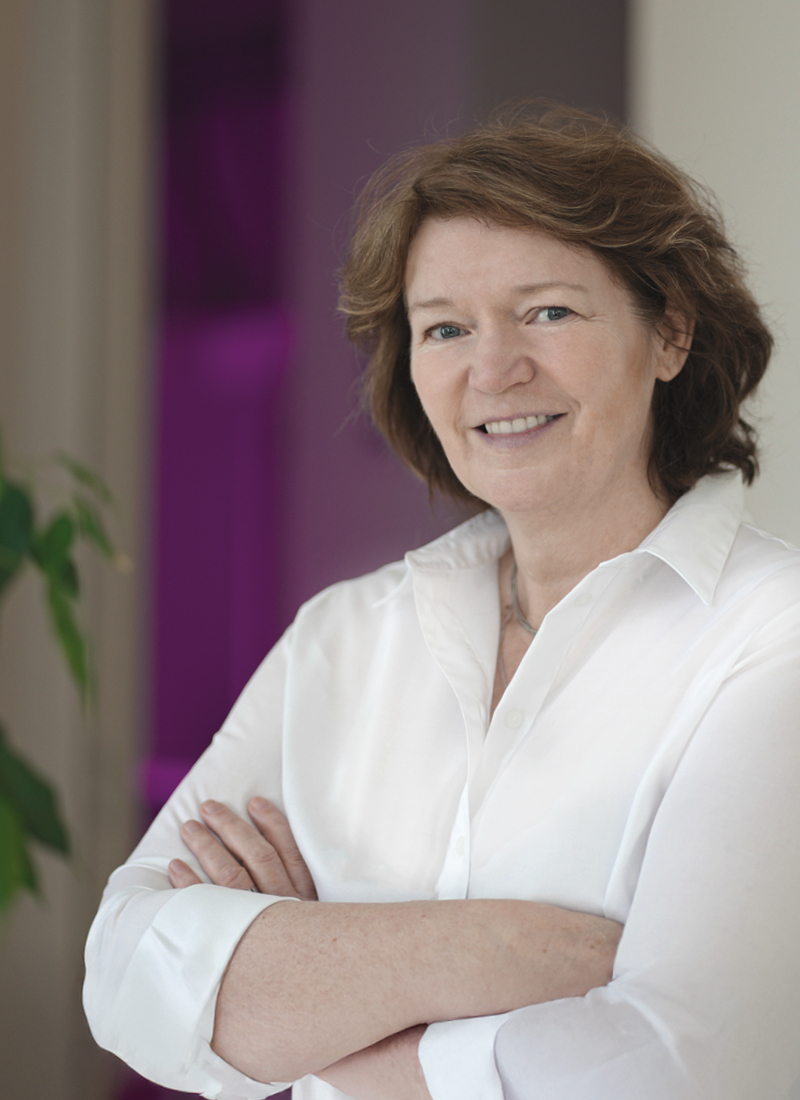 Portrait of a middle-aged woman with brown shoulder-length hair wearing a white blouse, smiling gently at the camera with her arms crossed; blurred purple shapes fill the background.